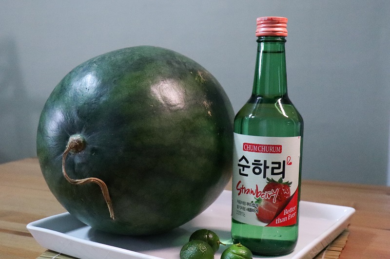 A watermelon, three limes and a bottle of strawberry soju displayed on a table.